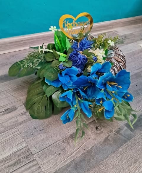 Decorative floral arrangement with blue flowers, green leaves, and a gold heart-shaped topper on wooden floor against turquoise wall