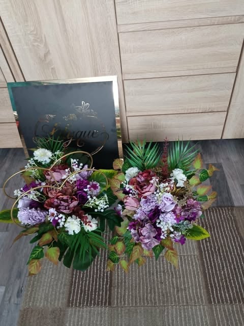 Floral arrangement with purple and white flowers, green palm fronds, and a dark chalkboard sign on wooden background
