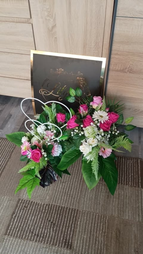 Floral arrangement with pink roses, white daisies, and green foliage in a black vase on a wooden floor with a chalkboard sign.