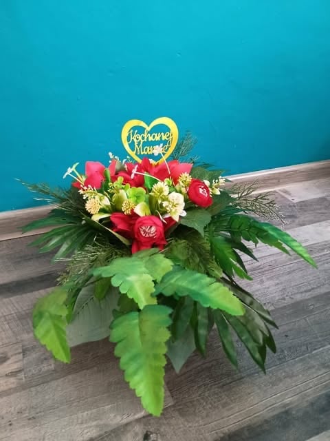 Floral arrangement with red and white flowers, green foliage, and a gold heart-shaped Je taime Maman topper on wooden surface against turquoise background