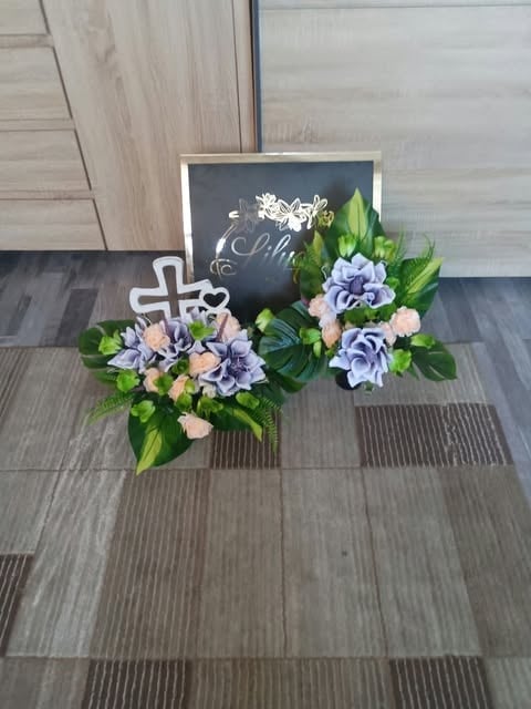 Floral arrangement with white cross and chalkboard sign on tiled floor, featuring blue and pink flowers with green foliage