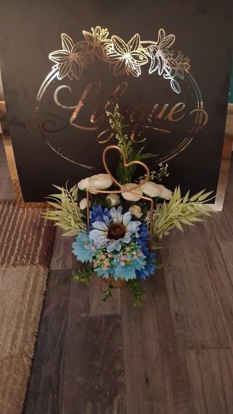 Decorative floral arrangement with blue and white flowers in a pot, placed in front of a black sign with gold text and flower designs.