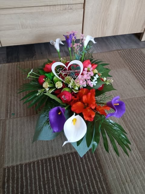 Decorative floral arrangement with red roses, purple irises, white calla lilies, pink flowers, green leaves, red ribbon bow, and a heart decoration on wooden flooring