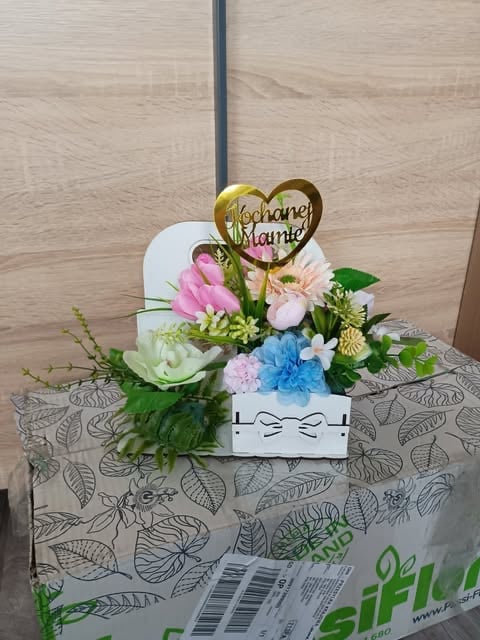 Floral arrangement with pink, white, and blue flowers in a white container topped with a gold heart charm reading Juste Marié on a patterned tablecloth