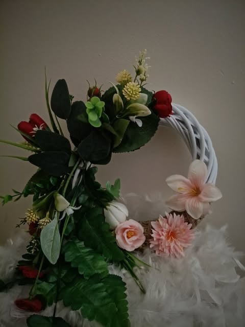 Decorative floral wreath with red roses, pink flowers, yellow blooms, green foliage, and white wicker base on neutral background
