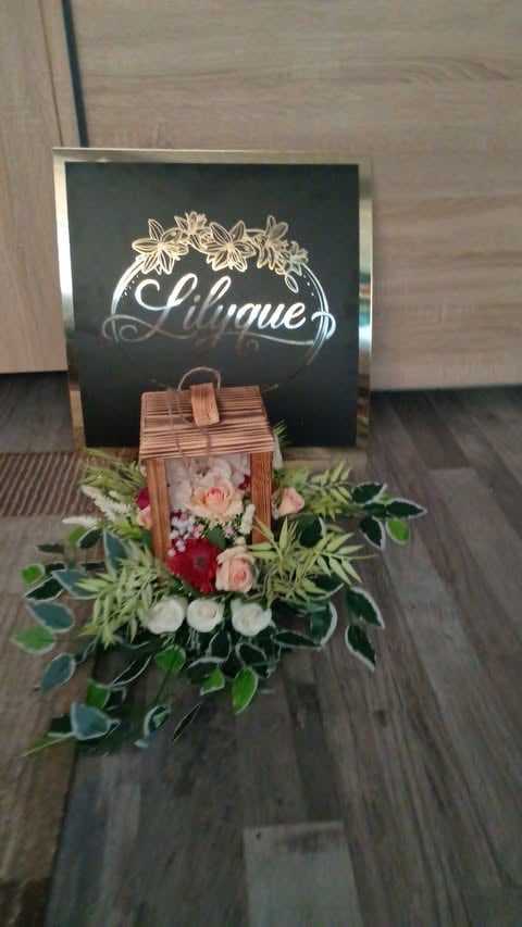 Wooden wicker basket arrangement with pink and red flowers and green foliage, displayed in front of a black sign reading Lilique with floral illustration