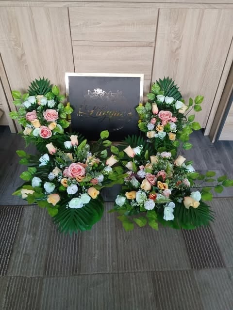 Floral arrangement display with pink, white, and peach flowers surrounding a black decorative sign on wooden flooring indoors