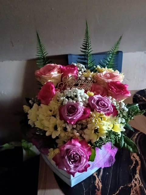 Colorful flower arrangement in light blue box with pink, yellow, and purple roses, carnations, greenery, and white babys breath