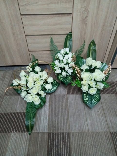 Three white flower arrangements with green foliage and fern leaves displayed on striped flooring against a wooden wall.