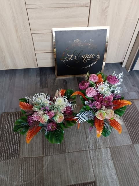 Floral arrangements with pink carnations, white chrysanthemums, and orange proteas displayed in front of a shop sign reading Libque on a dark floor.