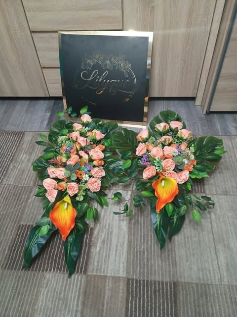 Elegant floral arrangement with pink roses and orange lilies in a butterfly shape, displayed with a black welcome sign on wooden flooring
