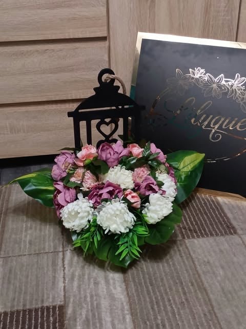 Floral arrangement with pink and white flowers, green leaves, black decorative lantern, and dark sign on wooden surface