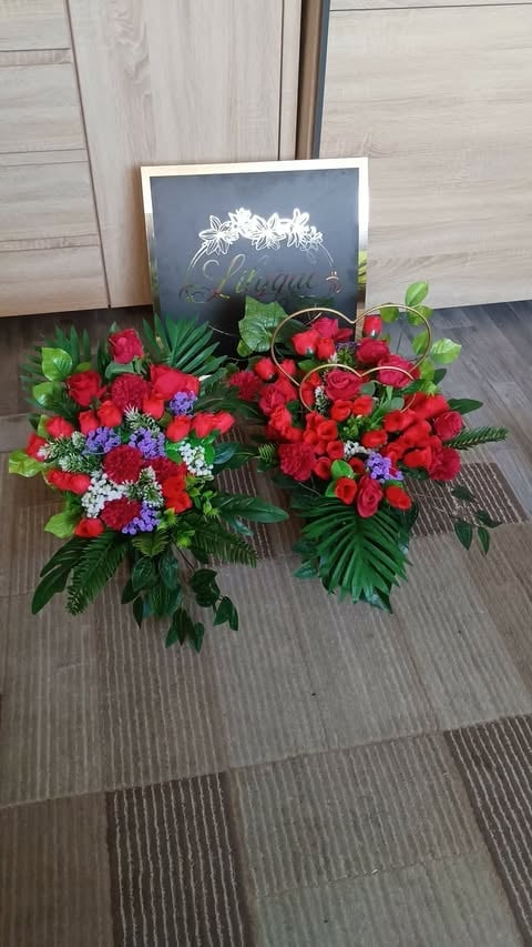Two red flower arrangements with purple accents and green foliage displayed on gray tile floor with a sign behind them