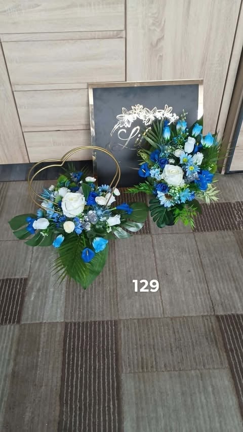 Floral wedding display with white roses, blue flowers, and green foliage arranged with a gold ring stand and chalkboard sign on wooden floor
