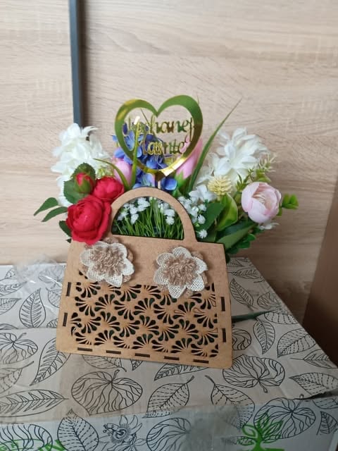 Decorative wooden flower arrangement with colorful blooms, white lace handles, heart-shaped sign, and patterned base on leaf-printed tablecloth