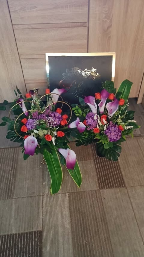 Colorful floral arrangement with pink calla lilies, purple hydrangeas, and orange roses on wooden floor with framed photo in background
