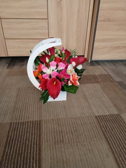 White curved floral basket with red anthuriums, pink roses, and mixed flowers on striped carpet near wooden doors
