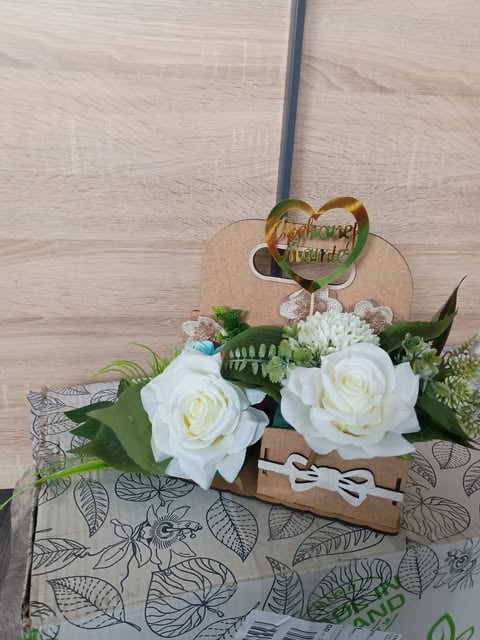 Wooden bride and groom sign on top of a floral wedding bouquet with white roses and greenery, displayed on a leaf-patterned cloth