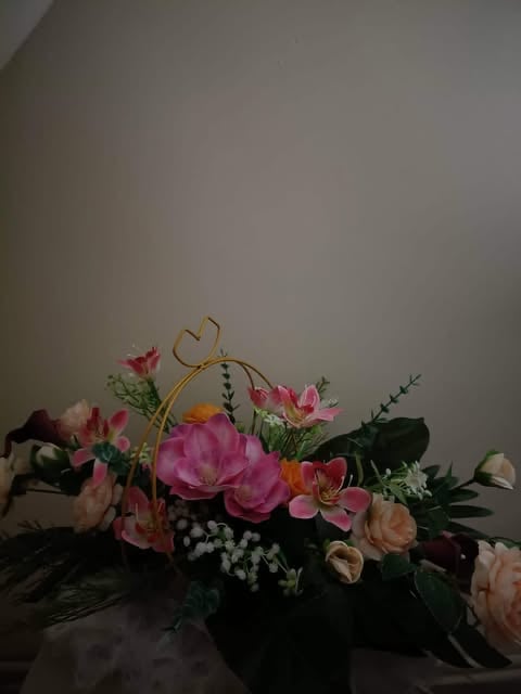 Floral arrangement with pink and peach flowers, greenery, and gold heart-shaped accent in black vase against white wall