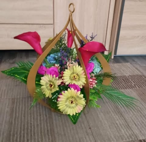 Hanging floral arrangement with yellow gerbera daisies, pink calla lilies, blue flowers, and green foliage in a gold teardrop-shaped basket with pink ribbon.