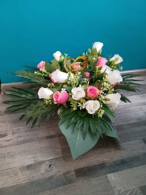 Floral arrangement in green vase with white, pink, and coral roses, greenery, and tropical foliage on wooden surface against teal wall