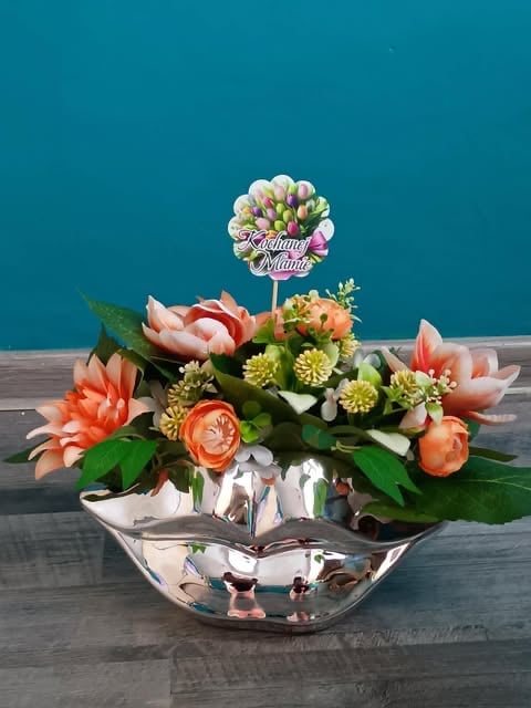Floral arrangement with coral and pink flowers in a silver bowl against turquoise background with Happy Monday sign