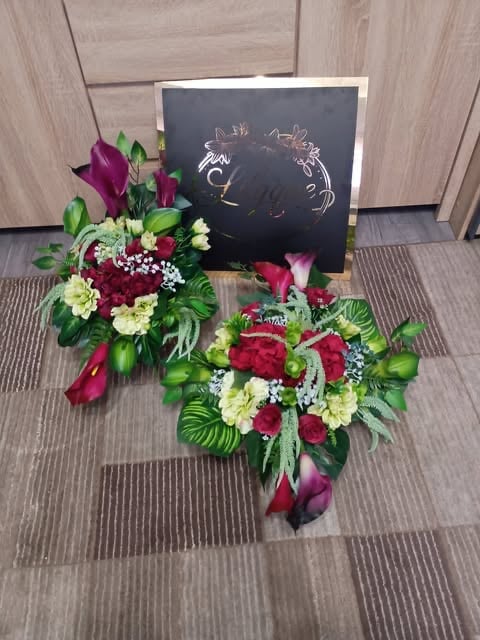 Two floral arrangements with burgundy roses, white flowers, and tropical leaves displayed on gray carpet next to a chalkboard sign