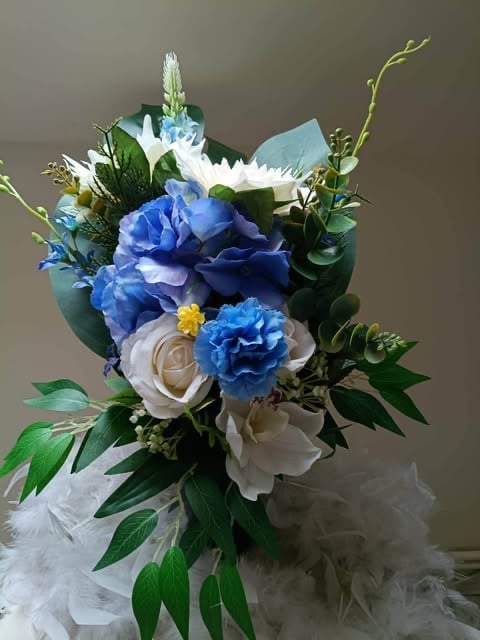 Elegant floral arrangement with blue and white flowers, greenery, and decorative elements in a tulle-wrapped vase against a neutral background