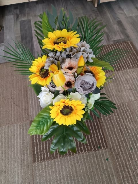 Vertical floral arrangement with yellow sunflowers, white roses, gray-purple flowers, and green palm leaves on textured floor
