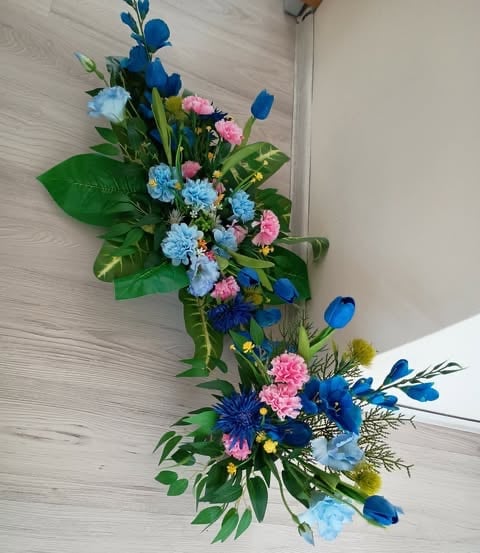Elegant floral arrangement with blue, pink, and light blue flowers with green foliage arranged vertically on a light wood wall