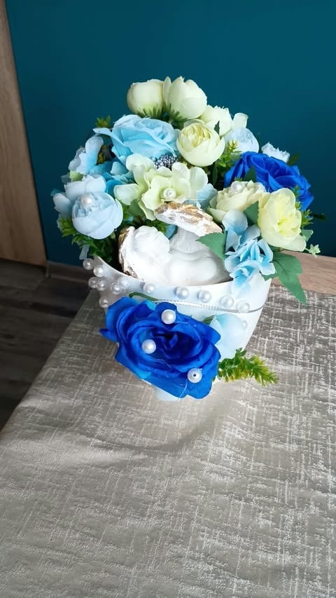 Artificial flower arrangement in white ceramic pot with blue, white, and cream flowers against teal background on gray surface