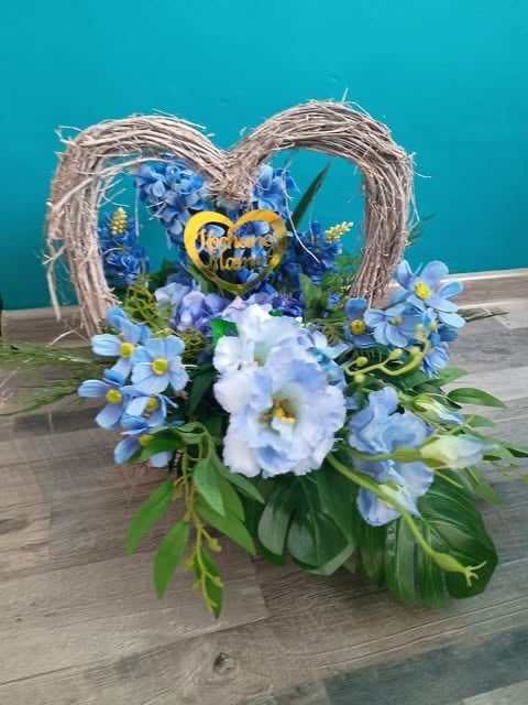 Decorative heart-shaped woven frame with blue and white flowers, golden heart ornament, and green foliage against turquoise background