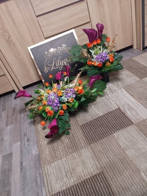 Colorful floral arrangement with purple calla lilies, orange roses, and purple flowers next to a black chalkboard sign reading Lilya on a wooden floor.