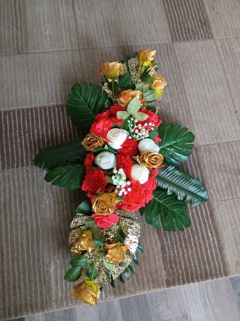 Floral cross arrangement with red carnations, gold roses, white flowers, and green tropical leaves on a textured gray background