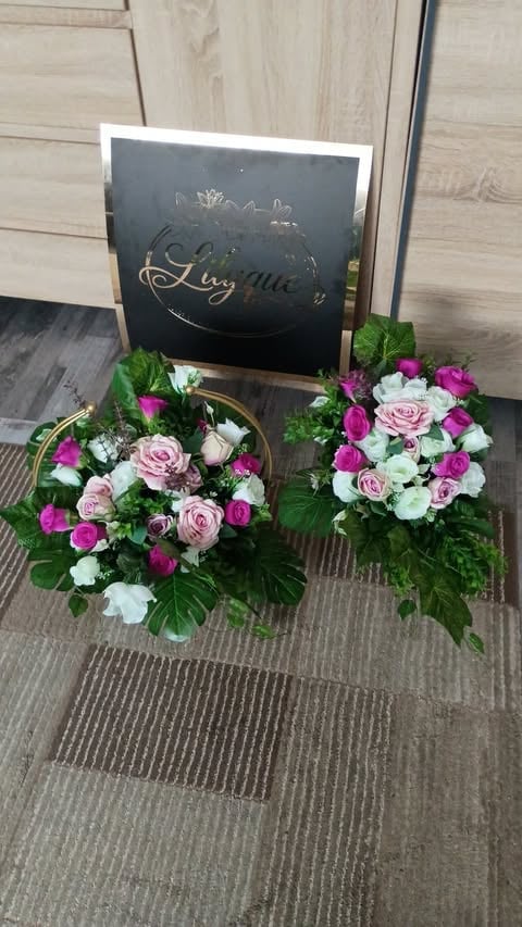 Two floral arrangements with pink roses, magenta carnations, and white flowers beside a black chalkboard sign on carpet flooring